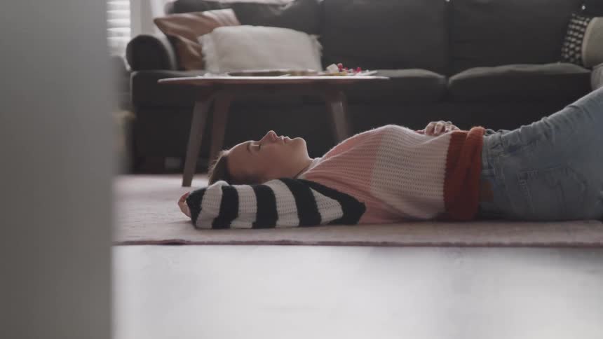 Woman with a sweater and jeans lies down on a carpet with eyes closed in the living room
