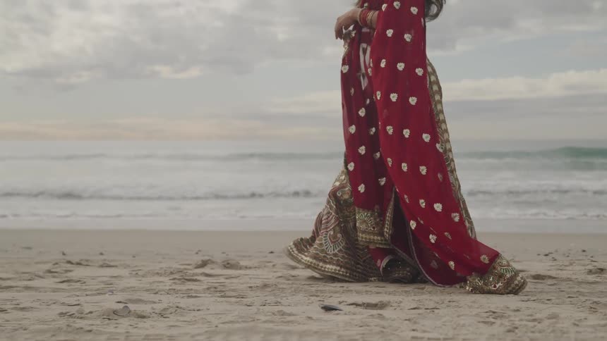 Woman with red traditional dress walks on white sand beach next to the ocean on a cloudy day