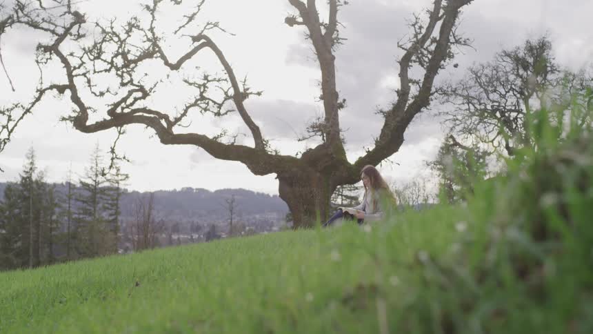 Woman with blonde hair sits on a green hill slope and reads a book on a cloudy day