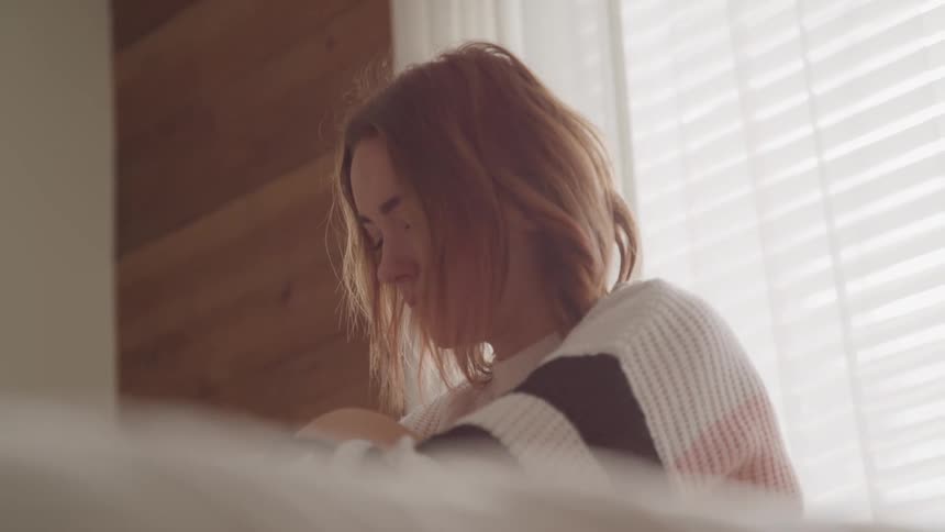 A young woman sits calmly in front of a window with the blinds drawn on a sunny day