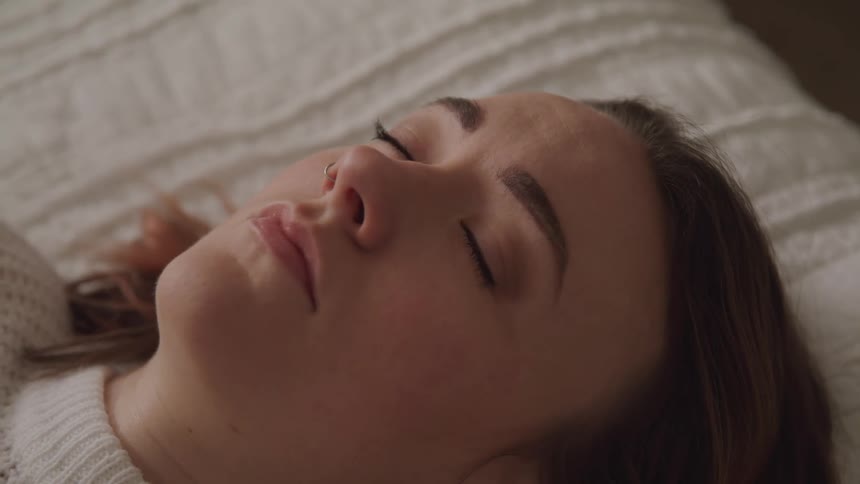 Woman with brown hair a white sweater and a nose ring sleeps on bed and opens her eyes