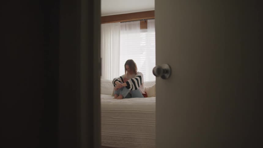 A woman sits on her bed with her arms around her knees and her head down