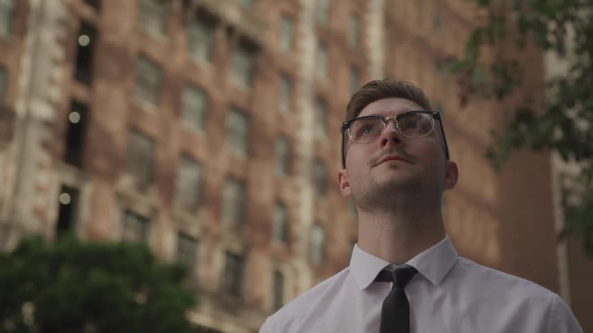 A man in a shirt and tie stands outside in a metropolis during the day and looks upwards at his surrounds