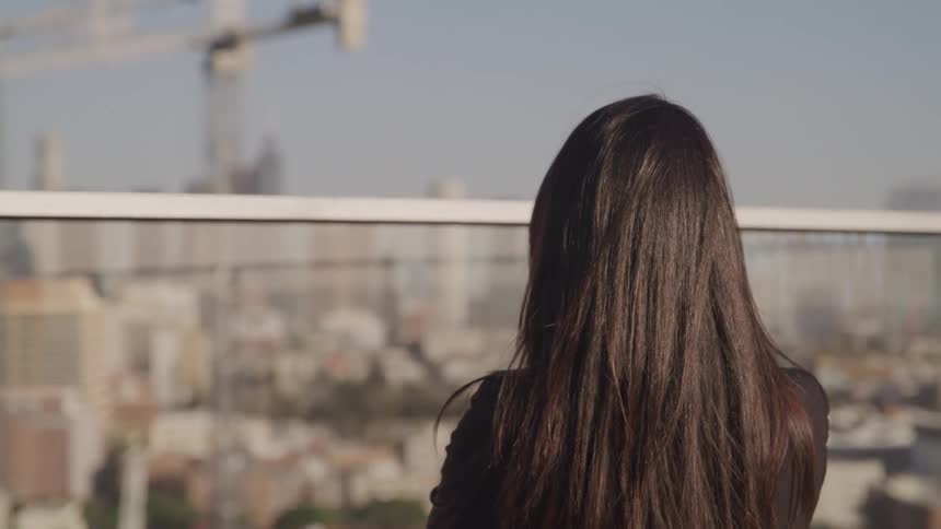 The wind blows the long brown hair of a woman on a balcony overlooking a city
