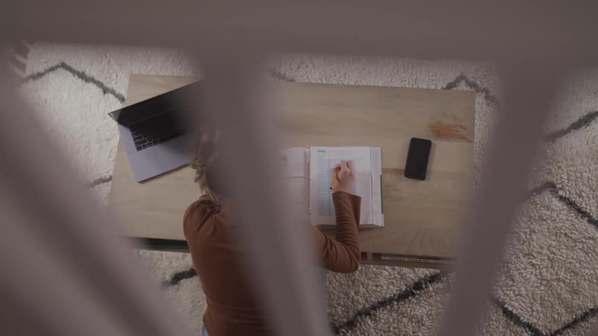Woman with a brown sweater jeans sits at a desk and studies from a book