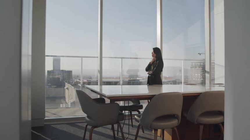 A middle aged asian woman in businesswear paces an empty conference room in a skyscraper