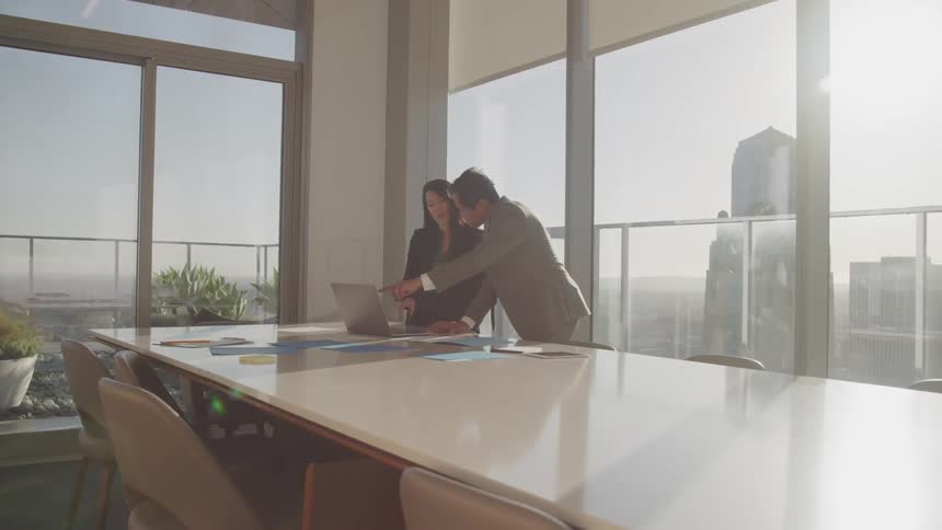 Man and woman look at a laptop sitting on a large table in a meeting room
