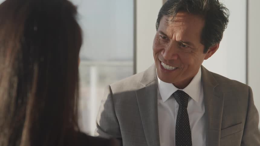 A man and woman in business wear shake hands while standing near a window in a skyscraper
