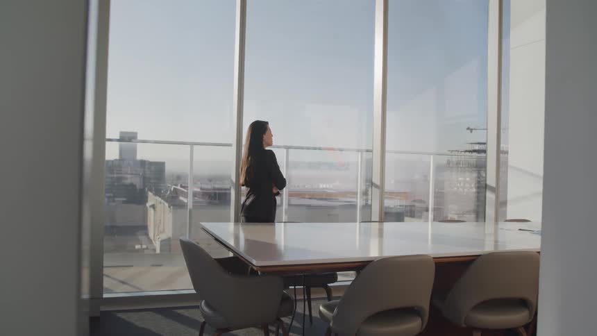Businesswoman with straight hair walks in the meeting room next to the office windows