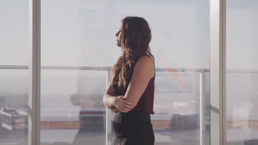 Businesswoman with wavy hair and a blouse and and a skirt walks and looks out the windows in the office