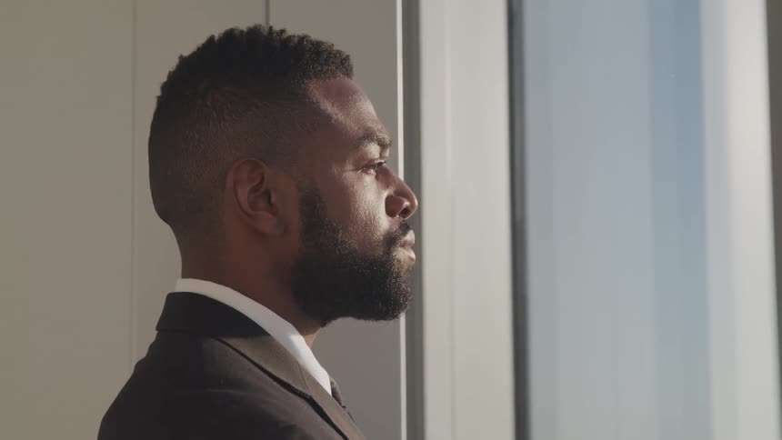 Side view of a black man with a beard and a suit looking out the window