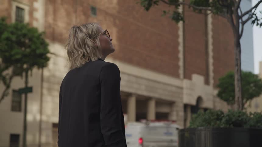 Blonde woman with glasses and a black blazer walks down the street in downtown area of a city
