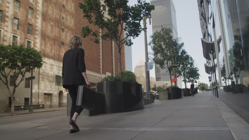 A woman carrying a briefcase looks up at the skyscraper next to her as she travels down a sidewalk in a metropolis during the day