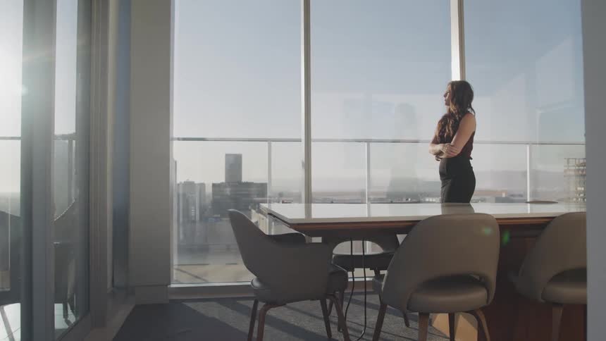 Businesswoman with wavy hair a black skirt and heels walks in the meeting room next to the office windows