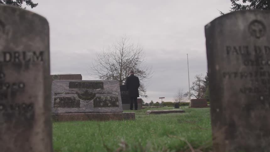 A man stands in front of a large headstone in a cemetery on a cloudy day