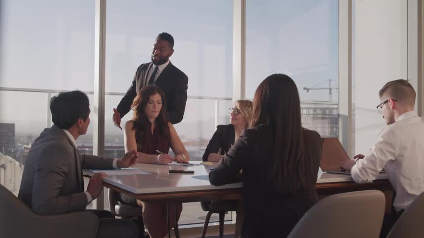 A man speaks as he walks around a conference table where several men and women sit