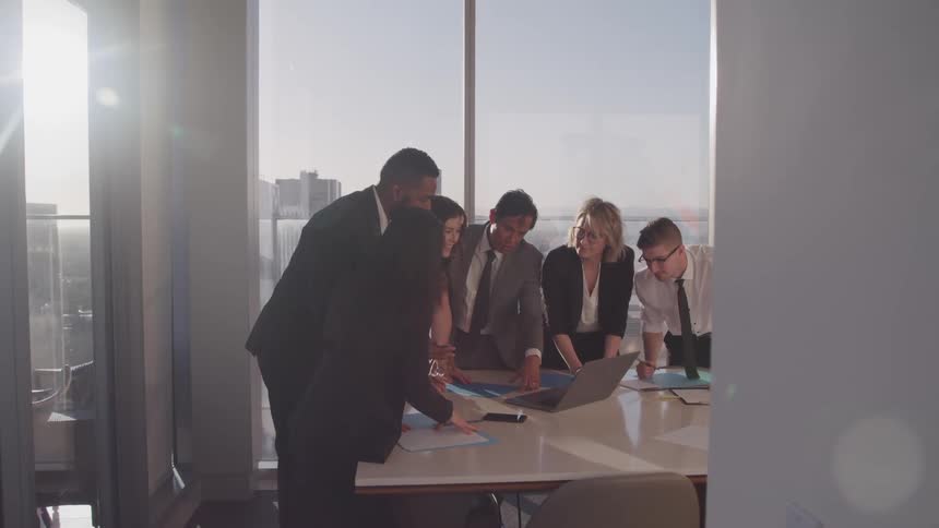 A group of people in an office gather around a laptop on a conference table