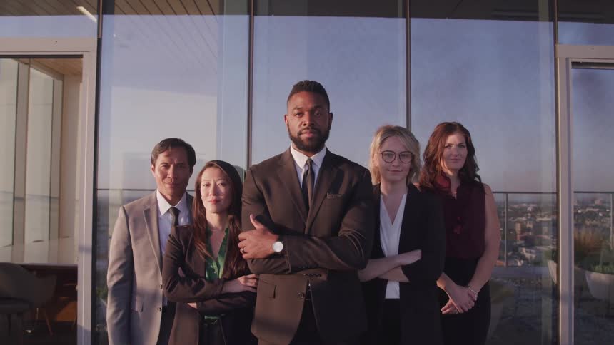 A group of five employees in suits pose in the office in front of a large window overlooking downtown