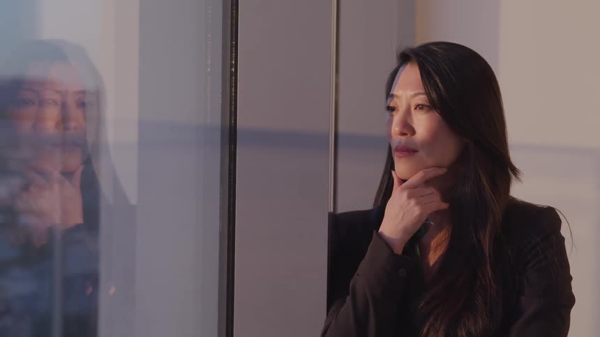 A middle aged asian woman cups her chin with one hand as she leans against the sill of a large window and gazes at the city on the other side