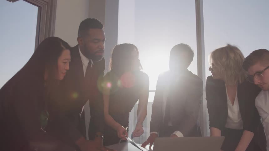 Business people huddle around laptop near window showing city discussing screen contents