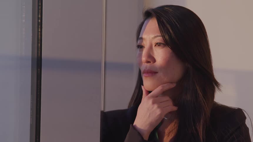 Asian businesswoman with black hair and a blazer holds her chin and looks out the office windows
