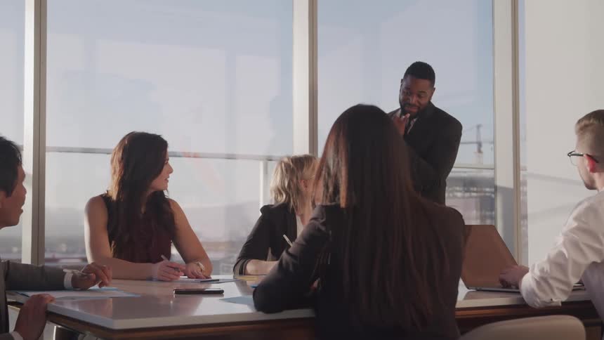 A man stands in an office and speaks to a group of men and women sitting around a conference table