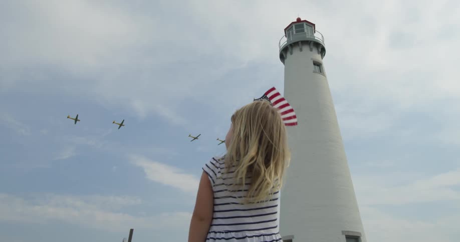 A girl holds up an american flag with planes and a lighthouse in the background