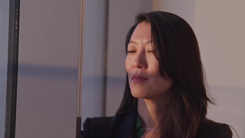 A middle aged asian woman looks out the window of an office before turning her head to look into the camera