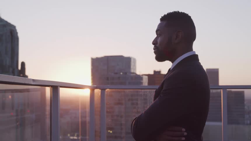 Side view of a businessman with a beard and a suit raising his heads on office balcony with tall buildings and sunset in the background