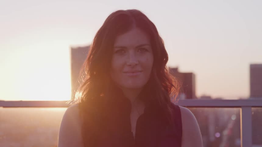 Businesswoman with black wavy hair stands on office balcony and faces the camera with eye contact at sunset