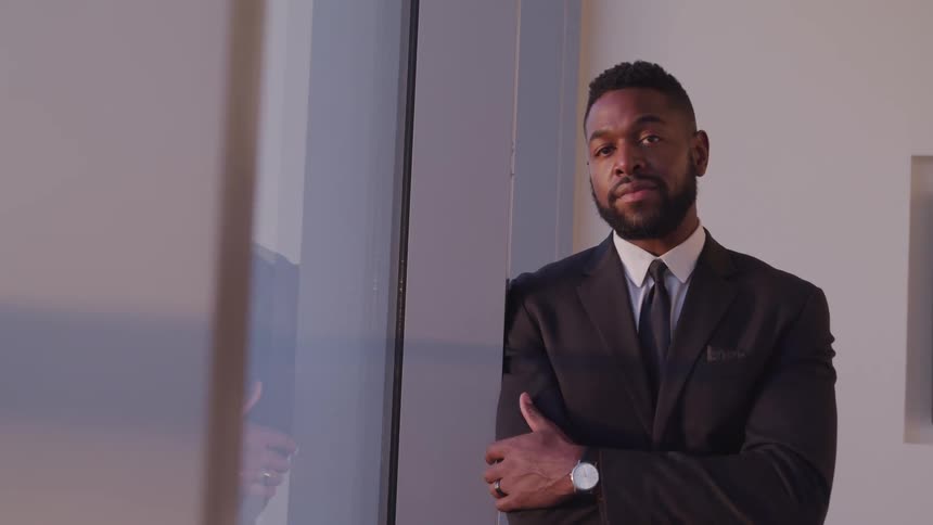 Black businessman with a beard and a suit leans against a wall and faces the camera with eye contact next to the office windows