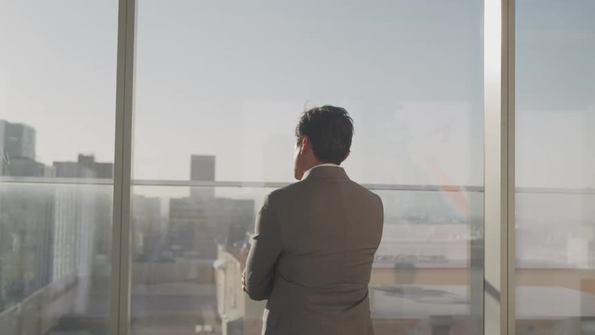A man in a suit standing by an office building window with buildings and the sky in the background