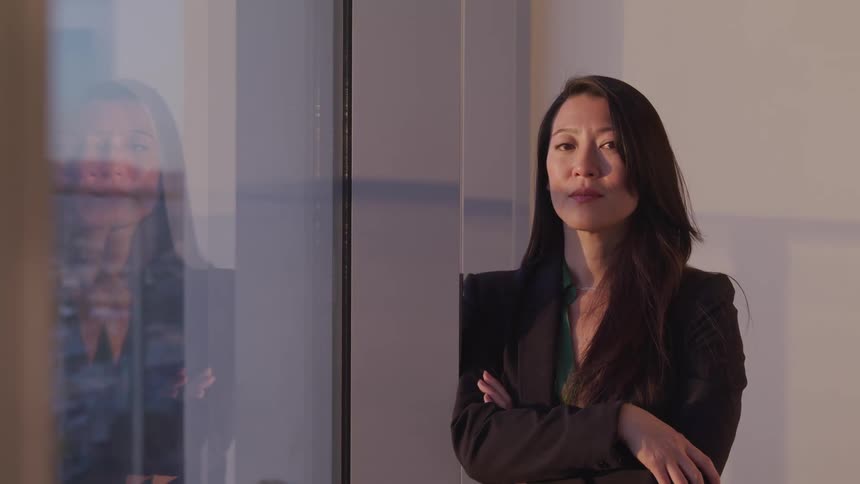Asian businesswoman leaning against a wall next to the office windows faces the camera with eye contact
