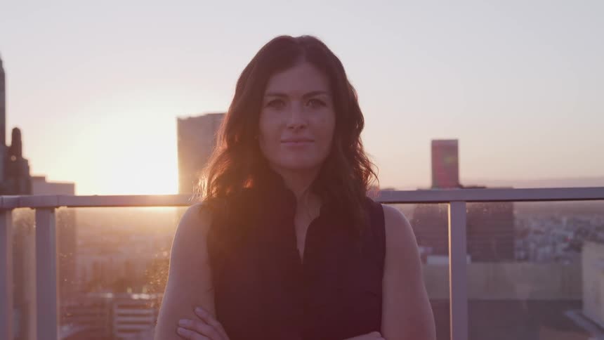 Businesswoman with black wavy hair stands on office balcony and faces the camera with eye contact at sunset