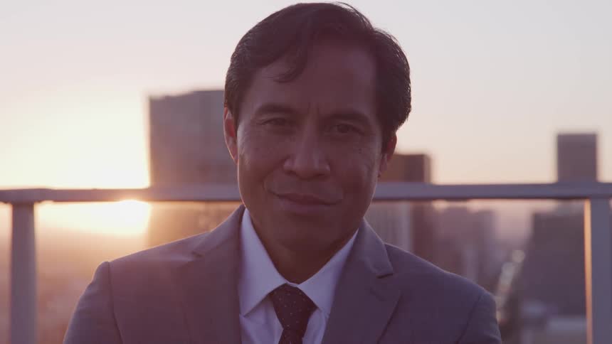 A man is standing on a balcony at dusk while wearing a suit