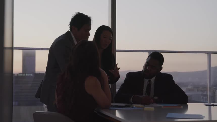 A group of people wearing business clothing gathered in an office in a skyscraper talk and write at a table