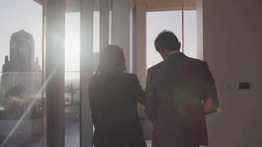 A man and woman walk together in an office room with large city view windows