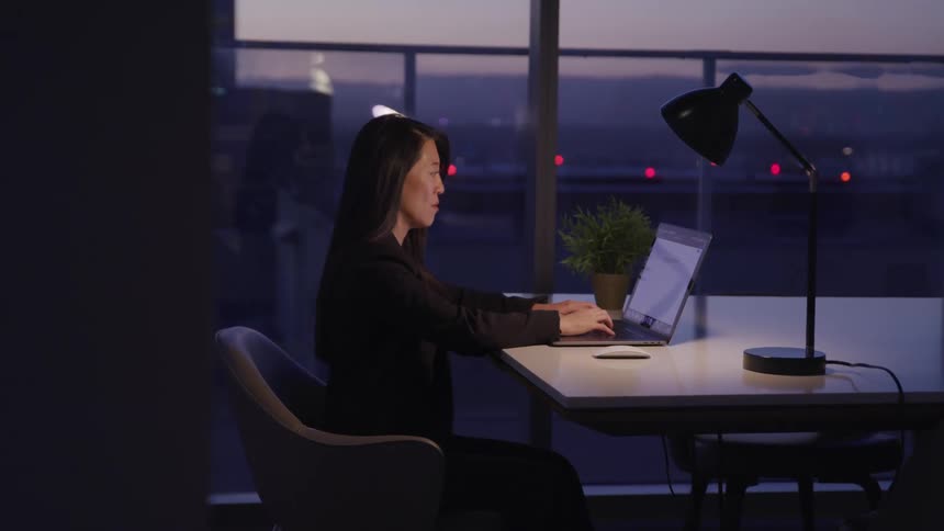 A middled aged asian woman types on a laptop while sitting at a table in an office in the late evening