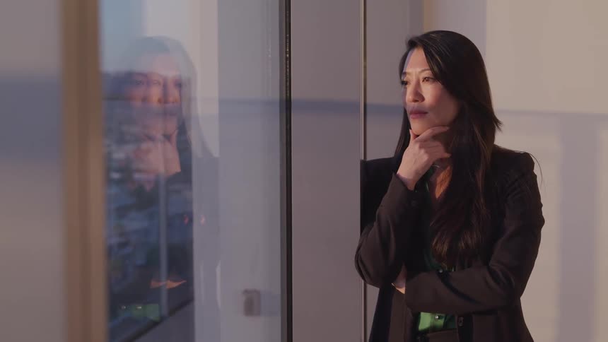 A woman in a business suit looks out the window in her office building as she has her hand propped under her chin