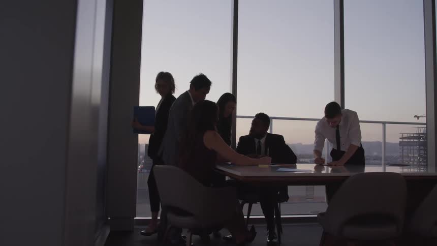 Business people conducting a meeting in a conference room are silhouetted against the sky at dusk