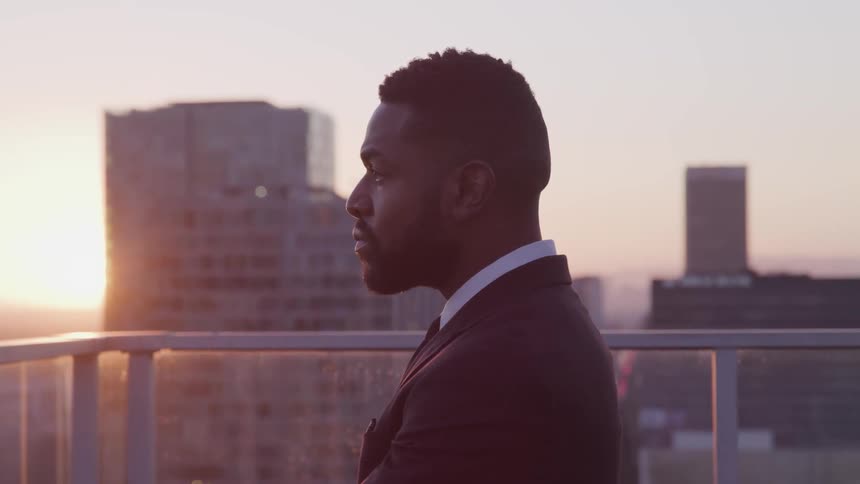 A man in a suit stands on a roof top and looks at the skyline