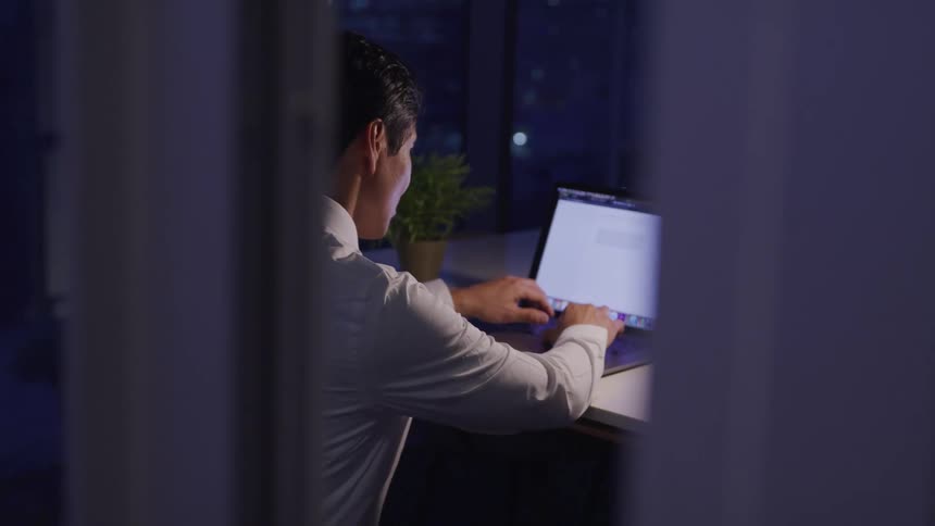 A man sitting at a desk typing on a laptop next to a lamp with windows in the background
