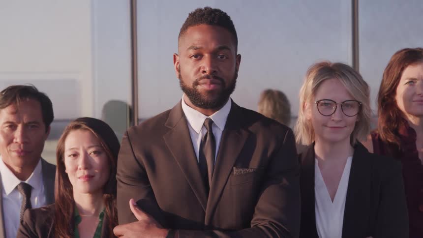 Five coworkers in business attire stand in a triangle formation and pose in front of a large office window