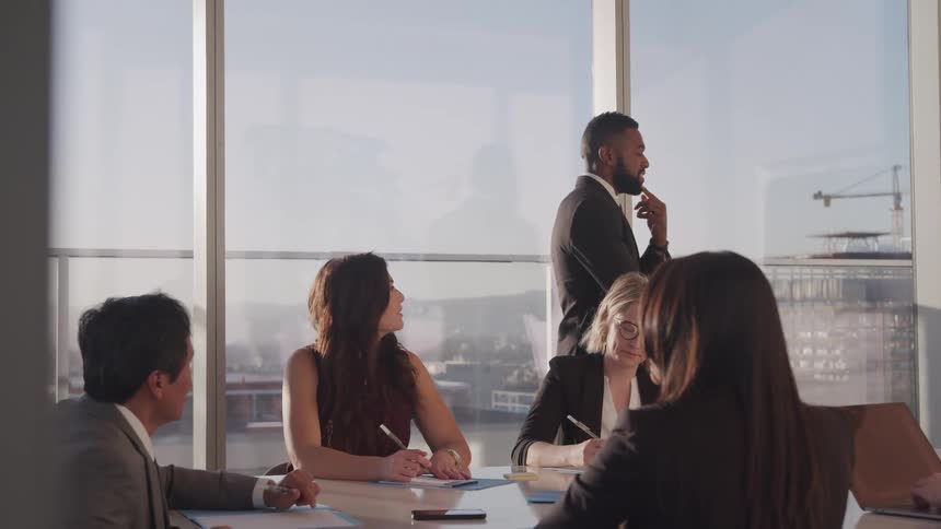 A group of people sit around a meeting desk and one man walks around the table