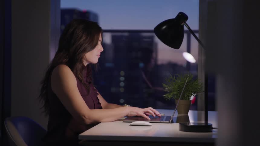 Businesswoman with wavy black hair sitting at a desk uses a laptop in the office at dusk