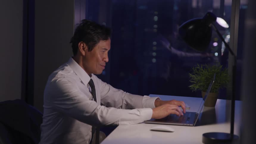 A man with a shirt and tie sitting at a desk typing on a laptop