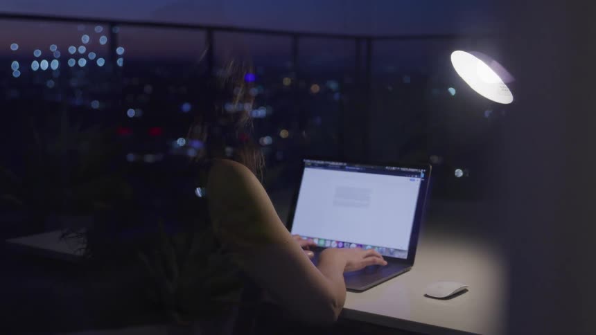 A persons reflection in the window is typing on a laptop