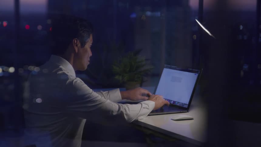 A man sitting in an office at nighttime works on composing a document on his laptop