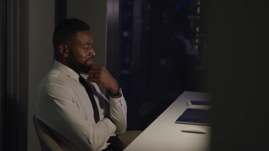 Black man sits and looks at a laptop on a table with a tablet sitting next to it