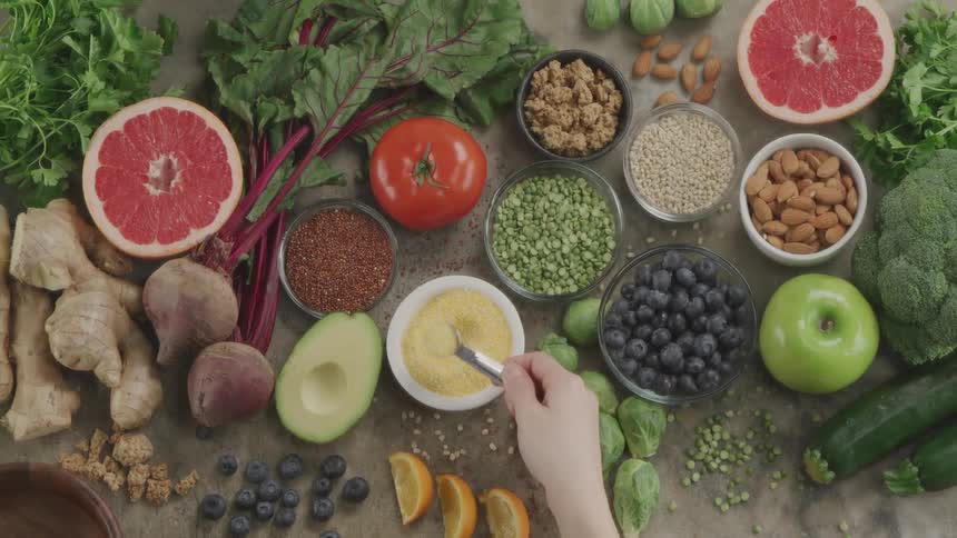 A person using a measuring spoon to scoop seeds from a white bowl surrounding by fruits and vegetables on a table